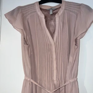 Ljusrosa kortklänning från H&M - Supersöt ljusrosa kortklänning från H&M med v-ringning, knyt i midjan och fina veckade detaljer framtill. Klänningen har korta, luftiga ärmar och är gjord i ett tunt, flowy material som känns lätt och somrigt. Fåtal använd. Skulle passa S-L