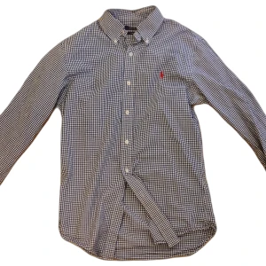 Rutig skjorta Polo Ralph Lauren S - Snygg blåvit rutig skjorta från Polo Ralph Lauren i slim fit-modell. Skjortan har klassisk krage, långa ärmar och knäppning framtill. Tillverkad i mjuk bomull och pryds av den ikoniska röda loggan broderad på bröstet.obs en knapp saknas