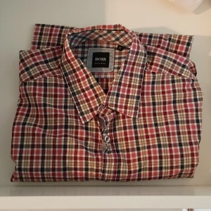 Rutig skjorta från Hugo Boss, M - Egentligen mer som nytt skick men satte den i mycket bra pga har ingen prislapp. Oanvänd. Snygg rutig skjorta från Hugo Boss i regular fit. Skjortan har klassisk krage, knäppning framtill och långa ärmar. Färgerna går i rött, blått, beige och vitt. Perfekt för dig som gillar en stilren look. 