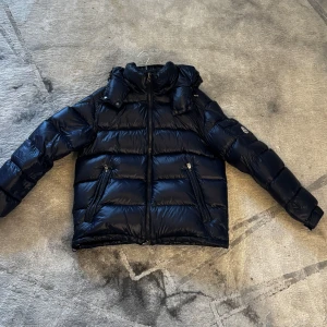 Svart Moncler pufferjacka - Säljer en navyblå Moncler Maya pufferjacka med glans, dragkedjor på fickorna och klassisk logga på ärmen. Jackan har hög krage och vadderad design som håller dig varm. Insidan har coolt Moncler tryck och innerficka med dragkedja. Helt ny, testat 1 gång bara. Size 2 Medium. Endast: 5150 kr