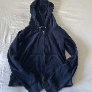 Mörkblå hoodie från Juicy Couture - Mysig mörkblå hoodie från Juicy Couture med dragkedja framtill, huva med snörning och klassiska kängurufickor. Liten broderad logga på bröstet. Perfekt för chill dagar eller när du vill vara extra comfy.