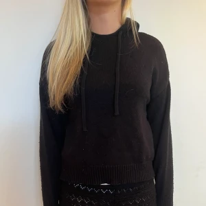 Svart stickad hoodie med snörning - Svart stickad hoodie med långa ärmar och snörning i halsen. Tröjan har ribbade muddar vid ärmslut och nederkant. 