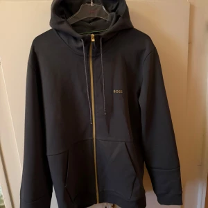 Svart hoodie från BOSS med gulddetalj och  dragkedja - Snygg svart hoodie från BOSS med guldfärgad dragkedja och diskret logga på bröstet. Tröjan har huva med snörning, långa ärmar och praktiska fickor framtill. Perfekt för en chill och stilren look.