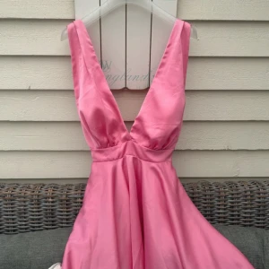 Rosa kortklänning med djup v-ringning - Supersöt rosa kortklänning i glansigt satinliknande material. Klänningen har djup v-ringning både fram och bak, breda axelband och en flowy kjol. Köpt i Santorini, ingen storlek men skulle säga att den är mellan S och M ☺️