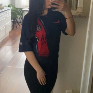 PSG fotbollströja Nike blå/röd - Säljer en Paris Saint-Germain fotbollströja från Nike. Tröjan är marinblå med en röd vertikal rand och vita detaljer, PSG-logga på bröstet och röd Nike-swoosh. Tillverkad i lätt och ventilerande material, perfekt för match eller träning.