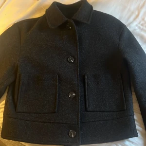 Mörkgrå kappa med stora fickor - Snygg svart jacka från lager 157. Med klassisk krage och stora framfickor. Jackan har raka linjer, knäppning med stora knappar och helsvart foder. Perfekt för dig som gillar minimalistisk och clean stil.