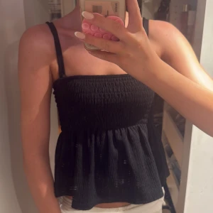 Svart smockad linnetopp med volang - Supersöt svart linnetopp med smockad överdel och volang nertill. Tunt och luftigt tyg med smala axelband, perfekt för varma dagar. Enkel att matcha med jeans eller kjol för en chill look. Snygg för vinter och sommar 