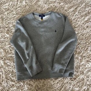 Grå sweatshirt från Polo Ralph Lauren - Klassisk grå sweatshirt från Polo Ralph Lauren med rund halsringning och broderad logga på bröstet. Tröjan har långa ärmar och ribbade muddar vid ärmslut och nederkant. Perfekt för en chill och stilren look.