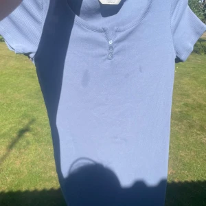 Ljusblå ribbad topp med knappar - En ljusblå ribbad klänning tait med korta ärmar och rund halsringning. Toppen har tre små knappar framtill som ger en chill och enkel look. Perfekt för dig som gillar basic men ändå vill ha en liten detalj. Skön och stretchig passform. Några fett flekar +frakt är redan tillagt