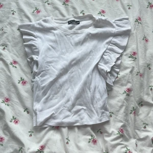 Vit volangtopp från Zara - Supersöt vit topp från Zara med volangdetaljer på axlarna. T-shirten är ribbad och perfekt för sommaren! 