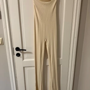 Beige byxdress i stretchigt material - Säljer en beige, ärmlös byxdress i mjukt och stretchigt material. Modellen är figurnära med långa ben och hög midja, perfekt för en clean och stilren look. Passar dig som gillar minimalistiska vibes och vill ha något bekvämt men ändå snyggt.