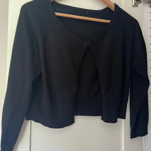 Svart bolero - En svart, enkel kofta med v-ringad hals och lång ärm. Modellen är croppad och har ingen synlig knäppning framtill, vilket ger en clean och stilren look. Perfekt att slänga över en topp eller klänning för en snygg layering-effekt. Passar mig som har S till xs. Säljer då den inte kommer till användning. Har en krok som sätts ihop