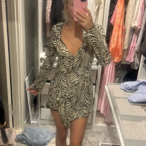 Zara klänning  - Kort zebramönstrad omlottklänning med v-ringning och långa ärmar. Klänningen har svartvita ränder och knyt i midjan för en snygg passform. Perfekt för dig som vill sticka ut med en trendig look.