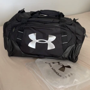 Svart 3.0 träningsväska från Under Armour. - Stor svart träningsväska från Under Armour med silvrig logga på framsidan. Väskan har flera fack med dragkedja, robusta handtag och justerbar axelrem. Perfekt för gymmet eller sportaktiviteter. Den är helt ny och oanvänd då jag köpte fel storlek.( 61L) 