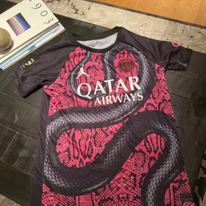 PSG Mbappé x Jordan ormskinnströja - Säljer en Paris Saint-Germain fotbollströja i samarbete med Jordan, med Mbappé 7 på ryggen. Tröjan är svart och rosa med ett unikt ormskinnsmönster och stor orm på framsidan. Qatar Airways sponsortryck och PSG-logga på bröstet. Tillverkad i lätt funktionsmaterial.