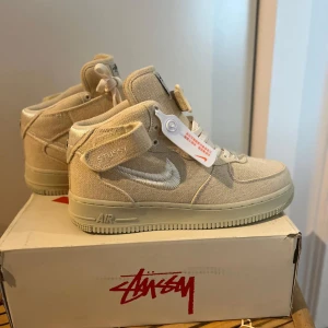 Nike Air Force 1 Stussy beige canvas deadstock  - Helt nya Nike Air Force 1 Stussy i beige canvas med vita detaljer och broderad Swoosh. Skorna har högt skaft, snörning och kardborreband vid ankeln. Stussy-logga på plösen och bak. Perfekta för dig som gillar clean och stilren streetwear.