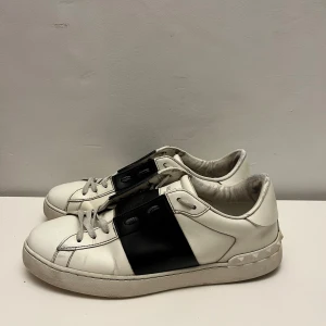 Valentino Garavani Open sneakers vit/svart - Snygga Valentino Garavani Open sneakers i vitt skinn med bred svart läderdetalj över mitten. Klassisk rund tå, platt sula och coola nitar baktill på sulan. Skorna har snörning och en chunky vibe som passar streetstyle. Märkeslogga på plösen och sula med rutmönster. Jättebra skick och 200 extra för tvättade innan jag skickar för maximal skick! Pris på tvätt 2200 och pris utan 2000