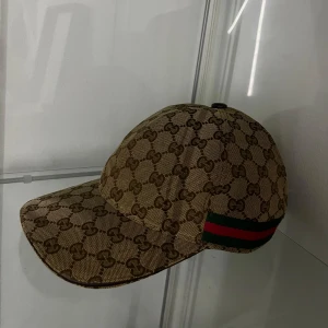 Gucci keps med GG-mönster - Snygg keps från Gucci i beige och brunt med klassiskt GG-monogram över hela kepsen. På sidan finns den ikoniska gröna och röda randen. Kepsen är tillverkad i bomull och polyester, perfekt för dig som vill sticka ut med en lyxig accessoar.