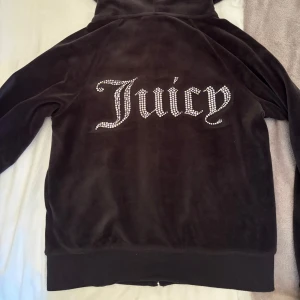 Juicy Couture velourjacka XS - Svart velour kofta luvtröja från Juicy Couture med huva, dragkedja och stora strasstenar som bildar texten 'Juicy' på ryggen. Jackan har långa ärmar, ribbade muddar och snörning i huvan. Klassisk logga framtill i strass. Perfekt för en chill och trendig look. Mycket bra skick.