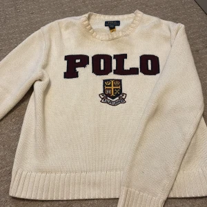 Vit stickad tröja Polo Ralph Lauren - Köpte för 1700kr på NK och säljer för mycket billigare☺️Vit stickad tröja från Polo Ralph Lauren med broderad logga och stort POLO-tryck i vinrött framtill. Klassisk rund halsringning och ribbade muddar. Perfekt för dig som gillar preppy college-stil.