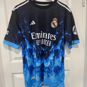 Real Madrid Mbappé #9 Adidas tröja - Snygg Real Madrid fotbollströja från Adidas med Mbappé och nummer 9 på ryggen. Tröjan har ett unikt blått och svart mönster, klubbmärke på bröstet och vita detaljer. Tillverkad i lätt funktionsmaterial med korta ärmar och tryckta sponsorer.