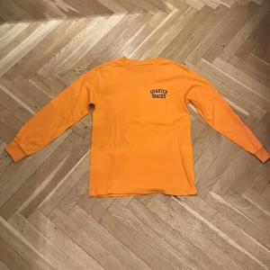 Orange långärmad tröja Quarter Snacks - Orange långärmad tröja från Quarter Snacks i 100% bomull. Framsidan har en liten svart logga på bröstet och baksidan har ett coolt tryck med tre grafiska bollar i olika mönster. Perfekt för dig som gillar streetwear och vill sticka ut.