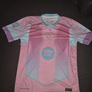 Barcelona Lamine Yamal fotbollströja - Säljer en Barcelona fotbollströja med Lamine Yamal och nummer 19 på ryggen. Tröjan är kortärmad och har en unik design i rosa, lila och ljusblå toner med klubbmärke och Spotify-logga framtill. Tillverkad i lätt och ventilerande Dri-FIT-material.