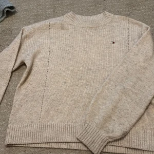 Beige stickad tröja Tommy Hilfiger - Beige stickad tröja från Tommy Hilfiger med diskret logga broderad på bröstet. Tröjan har ribbade muddar vid hals, ärmslut och nederkant samt långärmad passform. Perfekt för lager-på-lager och chill höst- och vinterstil.