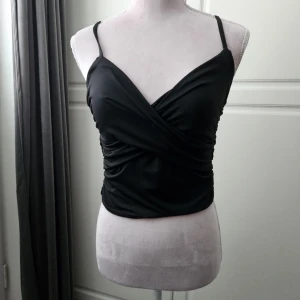 Svart omlott linnetopp med smala band - Snygg svart omlott linnetopp med smala axelband och rynkade sidor. Toppen har en croppad passform och djup v-ringning framtill. Perfekt för dig som gillar stilrena och trendiga plagg med en twist.