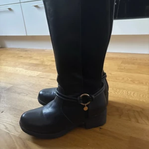 Svarta stövlar med gulddetalj - Snygga svarta stövlar i skinnimitation med rund tå och låg klack. Dekorativ guldring med hängande detalj runt ankeln och dragkedja på insidan för enkel påtagning. Perfekta till höst- och vinterlooken. Originalpris 1200kr