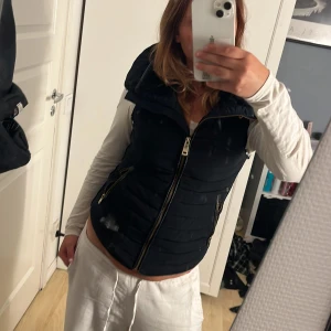 Svart dunväst från Zara Basic S - Svart quiltad dunväst från Zara Basic med dragkedja framtill och två fickor med dragkedja. Snygg hög krage och figurnära passform. Perfekt att slänga över en hoodie eller långärmad tröja för extra värme. Storlek S. Pris går att diskutera 💕💕