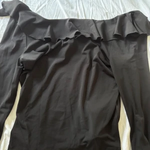 Svart offshoulder volangblus NA-KD - Svart blus från NA-KD i mjukt material. Toppen har långa ärmar med volangdetalj vid ärmslut och en bred volang längs axlarna. Perfekt för dig som gillar en trendig och feminin look med extra volym och snygg siluett. Aldrig använd