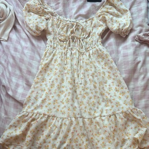 Blommig kortklänning från Gina Tricot - Supersöt kortklänning från Gina Tricot i ljus bas med små blommor i gult och rosa. Klänningen har puffärmar, smock i midjan, volangkant nertill och snörning framtill. Perfekt för sommarens alla tillfällen!