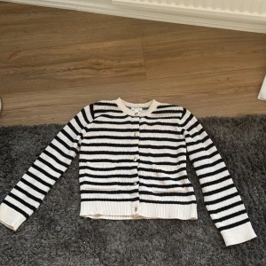 Randig svartvit kofta med knappar - Snygg stickad kofta i svart och vitt med horisontella ränder. Koftan har rund halsringning, långa ärmar och guldiga knappar framtill. Två små fickor framtill ger extra detalj. Perfekt för lager-på-lager och enkel att styla till jeans eller kjol.