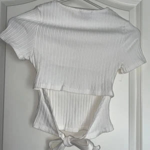 Vit ribbad topp med knytning - Säljer en vit ribbad topp med korta ärmar och snygg knytning i ryggen. Materialet är mjukt och stretchigt för extra komfort.