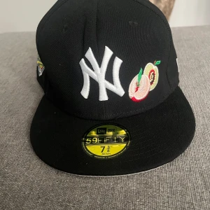New Era Yankees 59FIFTY keps svart - Svart 59FIFTY keps från New Era med New York Yankees-logga i vitt framtill, broderade frukter och färgglada detaljer. World Series-märke på sidan och MLB-logga bak. Klassisk flat peak och broderade ventilationshål. Material: polyester/ull.