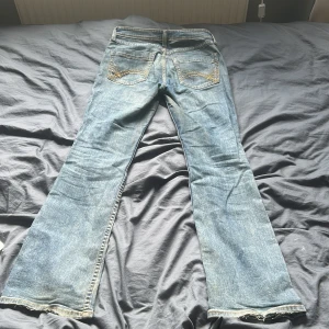 Blå bootcut jeans med broderade fickor - Snygga ljusblå jeans med bootcut passform och coola broderade detaljer på bakfickorna. Klassisk femficksmodell i jeansmaterial med slitna benslut för en avslappnad vibe. Perfekta för dig som gillar retrostil och vill sticka ut lite extra.