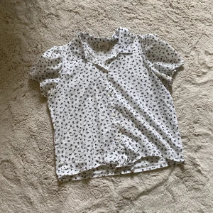 Vit blommig skjorta från KappAhl - Supersöt vit skjorta från KappAhl med svarta små blommor över hela tyget. Skjortan har kort puffärm, klassisk krage, knappar framtill och en snygg knytning längst ner. Perfekt för dig som gillar en fräsch och somrig look!