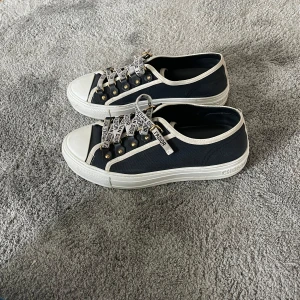 Christian Dior walk’n’dior skor  - Snygga låga sneakers från Dior i navy canvas med vita detaljer och vit sula. Skorna har unika snören med texten 'Christian Dior J'Adior' och guldfärgade metalldetaljer. Perfekt för dig som vill sticka ut med exklusiva och trendiga skor.
