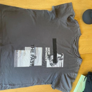 Stylish trendig Mörkgrå t-shirt med tryck - Snygg mörkgrå t-shirt med coolt grafiskt tryck framtill där det står 'THE PAST' och 'NEW MOMENT' i svart och vitt. T-shirten har rund halsringning och korta ärmar. Materialet känns mjukt och skönt, perfekt för en avslappnad stil.