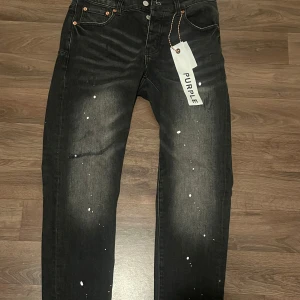 Svarta jeans från Purple Brand - Snygga svarta jeans från Purple Brand med coolt stänkmönster i vitt och slitna detaljer. Jeansen har klassisk femficksdesign, raka ben och knappgylf. Perfekta för dig som gillar streetwear och vill sticka ut lite extra.