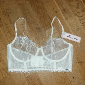 Vit spetsig korsettopp - Superfin vit korsettopp från Lindex, märket Ella M, med tunna axelband och transparent spets med broderade blommor. Toppen har bygel och dekorativ kant nertill, perfekt för att ge en snygg siluett. Säljs med originaltagg kvar. Stilren och romantisk vibe!