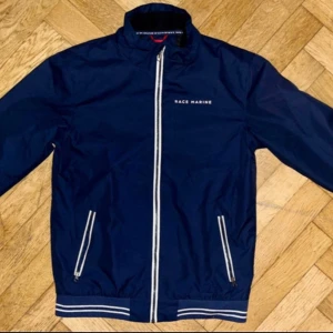 Marinblå vindjacka från Race Marine - Snygg marinblå vindjacka från Race Marine med vita detaljer och dragkedja framtill. Jackan har ståkrage, långa ärmar och två fickor med dragkedja. Ribbade muddar med vita ränder ger en sportig look. Perfekt för blåsiga dagar.