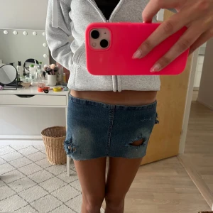 Jeansminikjol med slitningar och blommor - Superkort jeanskjol i blå denim med rå kant, slitna detaljer och hål framtill. På baksidan finns rosa och lila blommor med paljetter. Midjemått 44!!