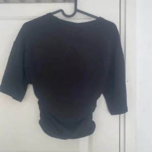 Svart topp med snörning i sidan - En svart topp med korta ärmar och cool snörningsdetalj i sidan som ger en snygg rynkad effekt. Perfekt för dig som gillar en enkel men ändå trendig look. T-shirten har en klassisk rund halsringning och är lätt att matcha med allt.