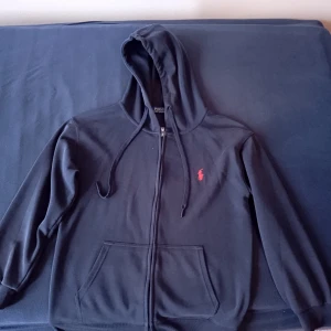 Mörkblå hoodie från Polo Ralph Lauren - Snygg mörkblå hoodie från Polo Ralph Lauren med dragkedja, huva med snörning och klassisk röd logga på bröstet. Tillverkad i 100% bomull och har känguruficka framtill. Perfekt för chill dagar och enkel att matcha med jeans eller joggers. Mer detaljer och bilder kan du få genom att skicka till mig privat. Pris kan diskuteras!!