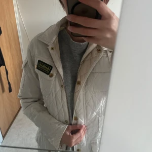 Vit quiltad jacka från Barbour - Säljer en snygg vit quiltad jacka från Barbour International. Jackan har guldiga knappar, dragkedja och coolt patch på bröstet. Klassisk krage och flera fickor framtill. Perfekt för dig som gillar stilren och tidlös design. 