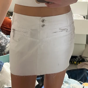 Vit kortkjol med rå kant och fickor - Vit kortkjol i bomull med rå, fransig kant nertill. Kjolen har dubbla knappar framtill, två dragkedjefickor fram och två bakfickor med lock och knapp. Enkel och clean design, perfekt till sommaren.