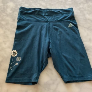 Träningsshorts i cykelbyxmodell - Jättefina träningsshorts ifrån Adidas i storlek s. Färgen är en mörk turkos färg (inte lika mörkblå som den ser ut på bild). Shortsen har en liten adidas logga och en blomdesign på sidan av shortsen. Perfekt för löpning eller annan aktivitet ☀️