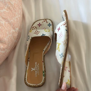 Louis Vuitton sandaler multicolor strl 39 - Snygga Louis Vuitton-sandaler i vitt skinn med det ikoniska monogrammönstret i flera färger (Multicolore). Klassisk modell med fyrkantig tå, platt sula och bred rem över foten – både bekväma och stilrena. Perfekta sommarskor som ger en lyxig touch till vilken outfit som helst. Denna design tillhör den populära Multicolore-kollektionen som togs fram i samarbete med Takashi Murakami, och som slutade produceras år 2015 – vilket gör dem eftertraktade som vintage. Pris kan diskuteras.
